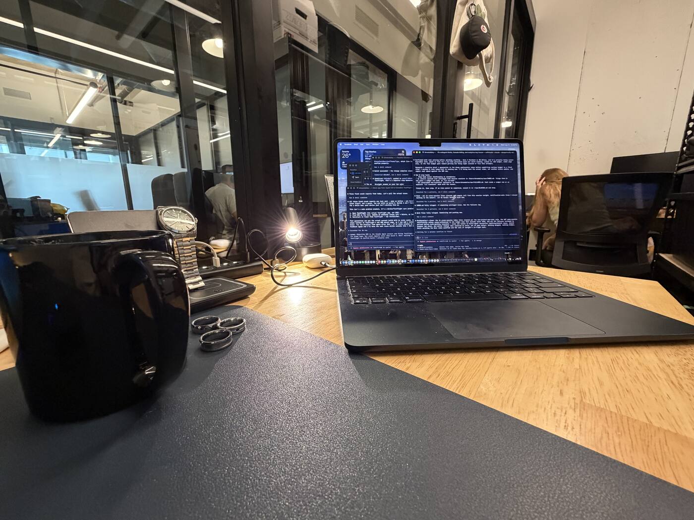 A bench desk after dark: an open laptop filled with terminal windows, a desk mat, a watch on a stand and a plain black mug, with the office floor and its glass partitions lit behind it and somebody still working at a monitor.