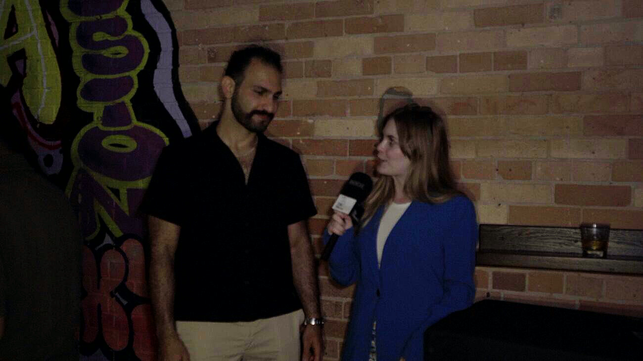 A TNAADO reporter in a blue blazer holding a branded microphone, interviewing a man against a brick wall with a painted mural, a second crew member at the edge of frame.