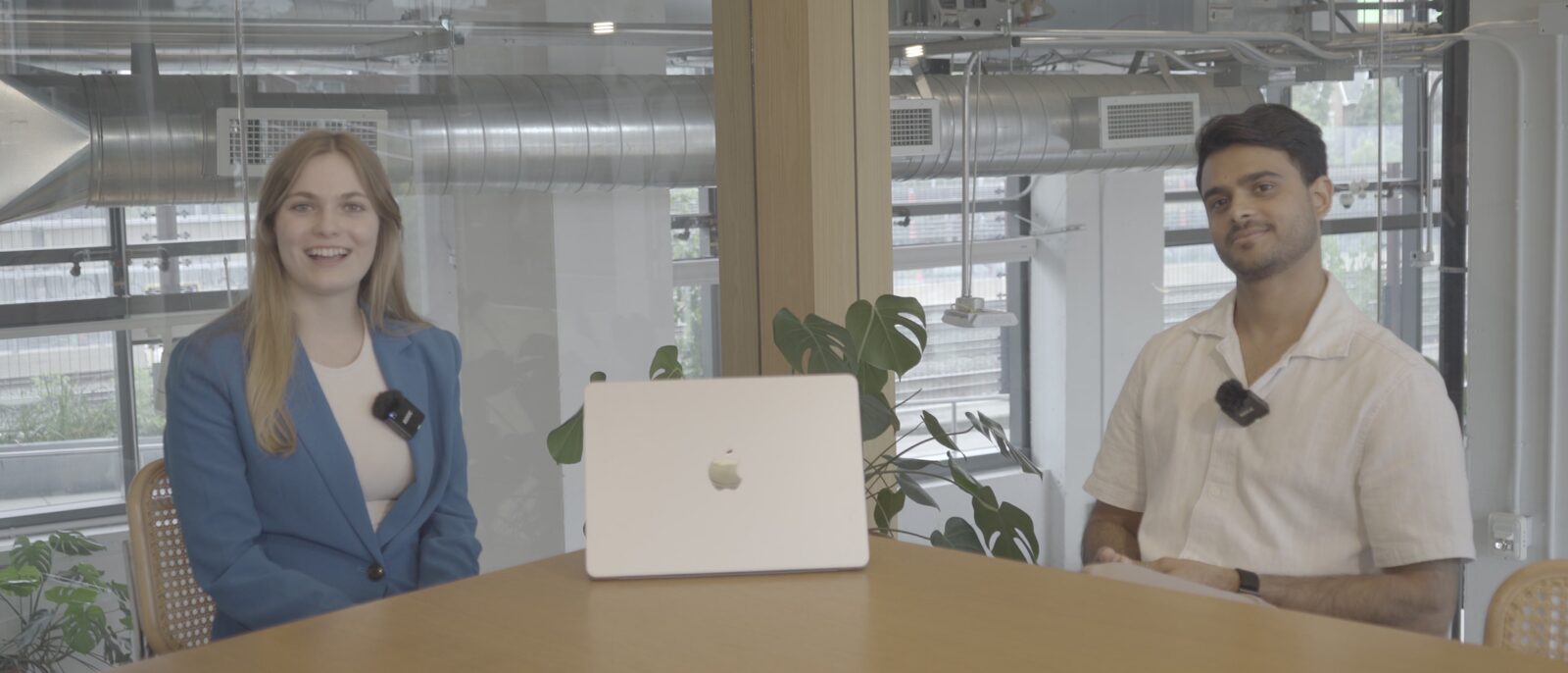 A finished interview two-shot: two people either side of a laptop at a wood table, both wearing lavalier microphones, in a bright glass-walled office.