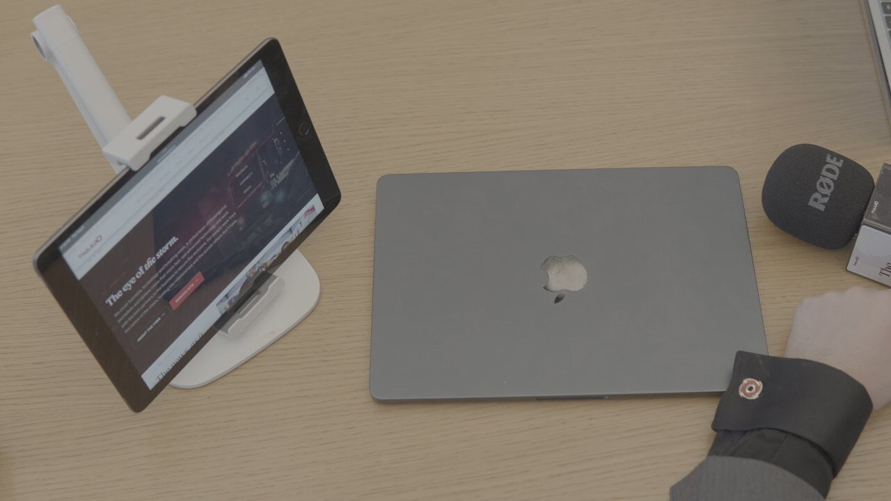 An overhead view of a pale wood table: an iPad in a stand showing the live tnaado.ca homepage, a closed laptop, and a microphone with a foam windshield.