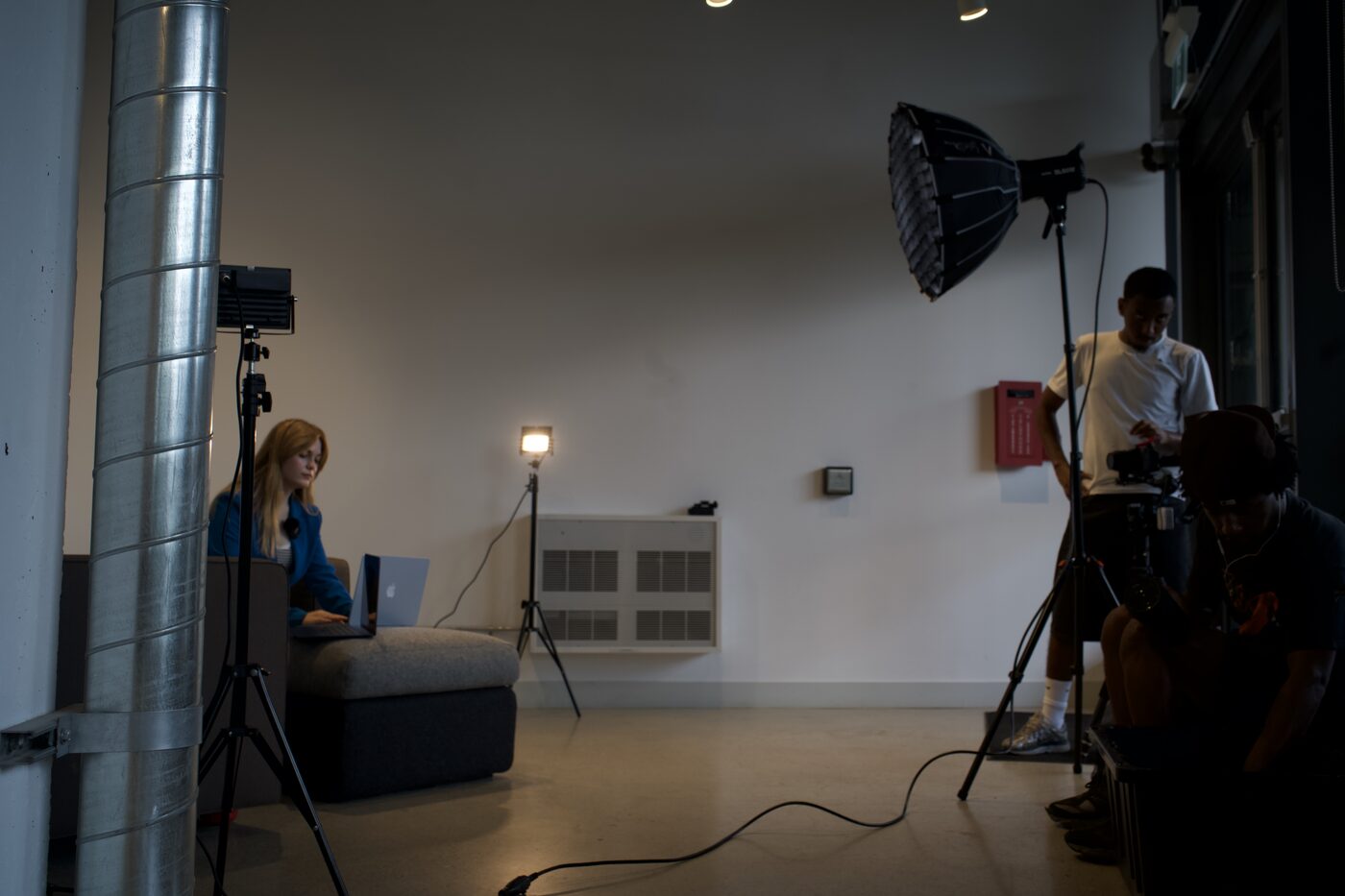 A lit interview set: a reporter working on a laptop at the left, LED panels on stands, and a camera operator and crew member under a large softbox at the right.