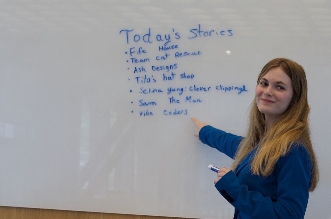A woman in a blue blazer stands at a whiteboard holding a marker and pointing at a handwritten list headed "Today's Stories": Fife House, Team cat Rescue, Ash Designs, Tito's hat shop, Selina Yung Clever Clipping, Sam The Man, Vibe Coders.