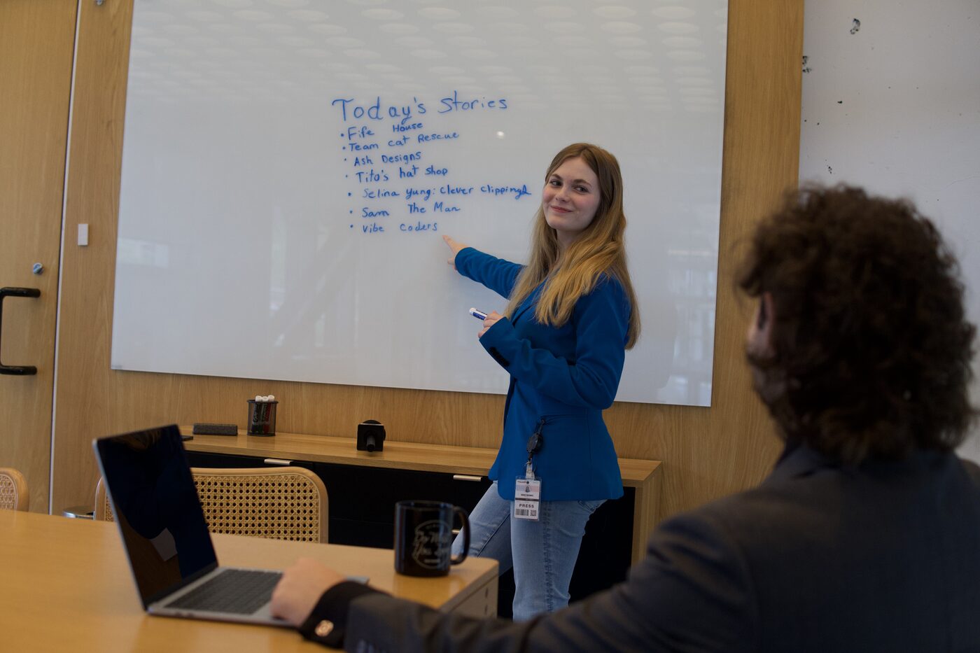 A reporter in a blue blazer stands at a whiteboard headed "Today's Stories", pointing at the last line of a seven-item list, with a colleague seated across the table.