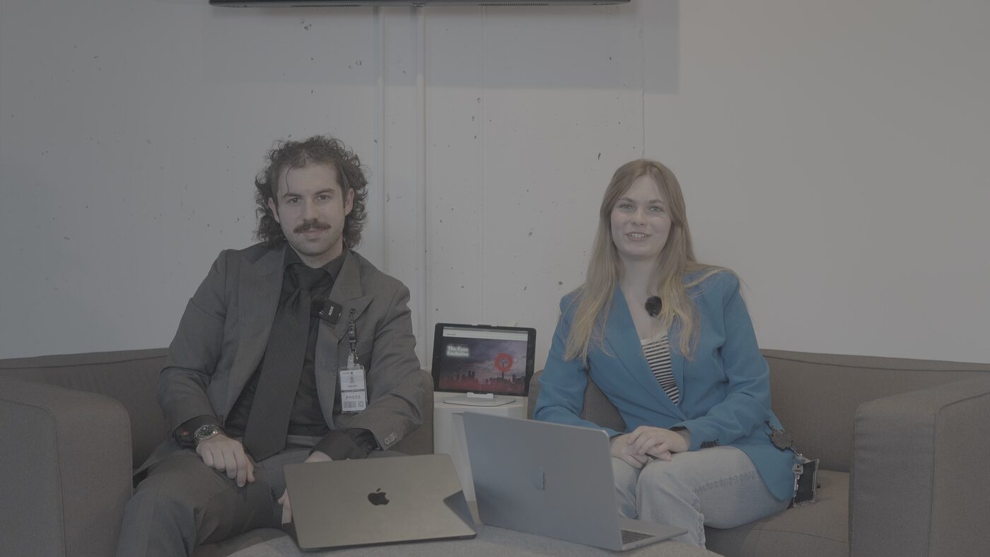 Two TNAADO presenters on camera at an interview setup, both wearing lavalier microphones, with a tablet showing the published page on a stand between them.