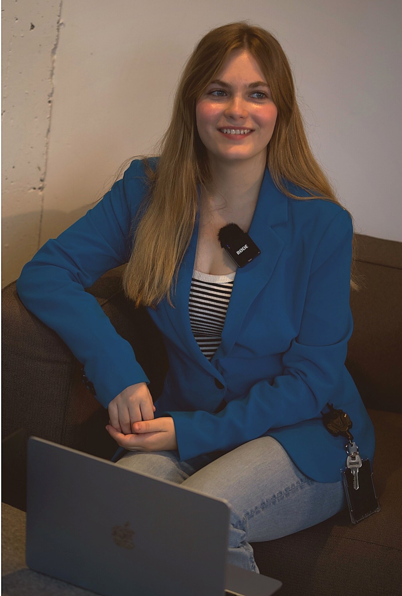 A reporter in a blue blazer with a RODE lavalier microphone clipped on, seated on camera with an open laptop beside her.