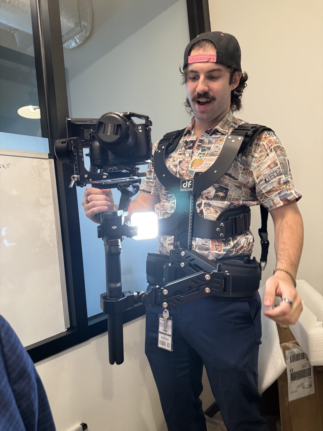 A videographer wearing a body-support vest and spring arm, holding a gimbal with a mirrorless camera and a small LED panel, a PRESS badge at his belt.