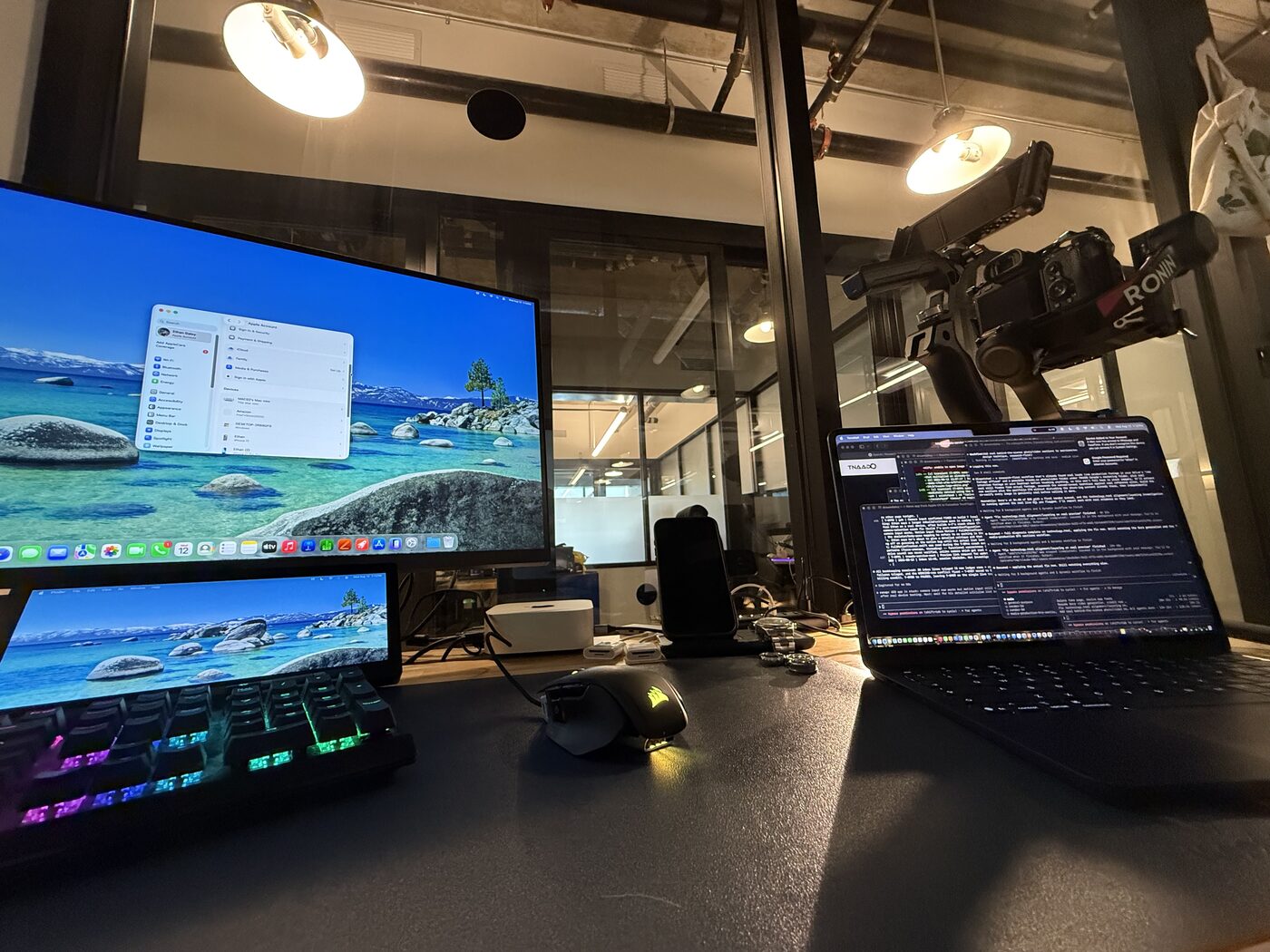 A desk in a Toronto office: dual displays and a backlit mechanical keyboard at the left, a laptop running a live coding session at the right, and a gimbal with a camera mounted suspended above it.