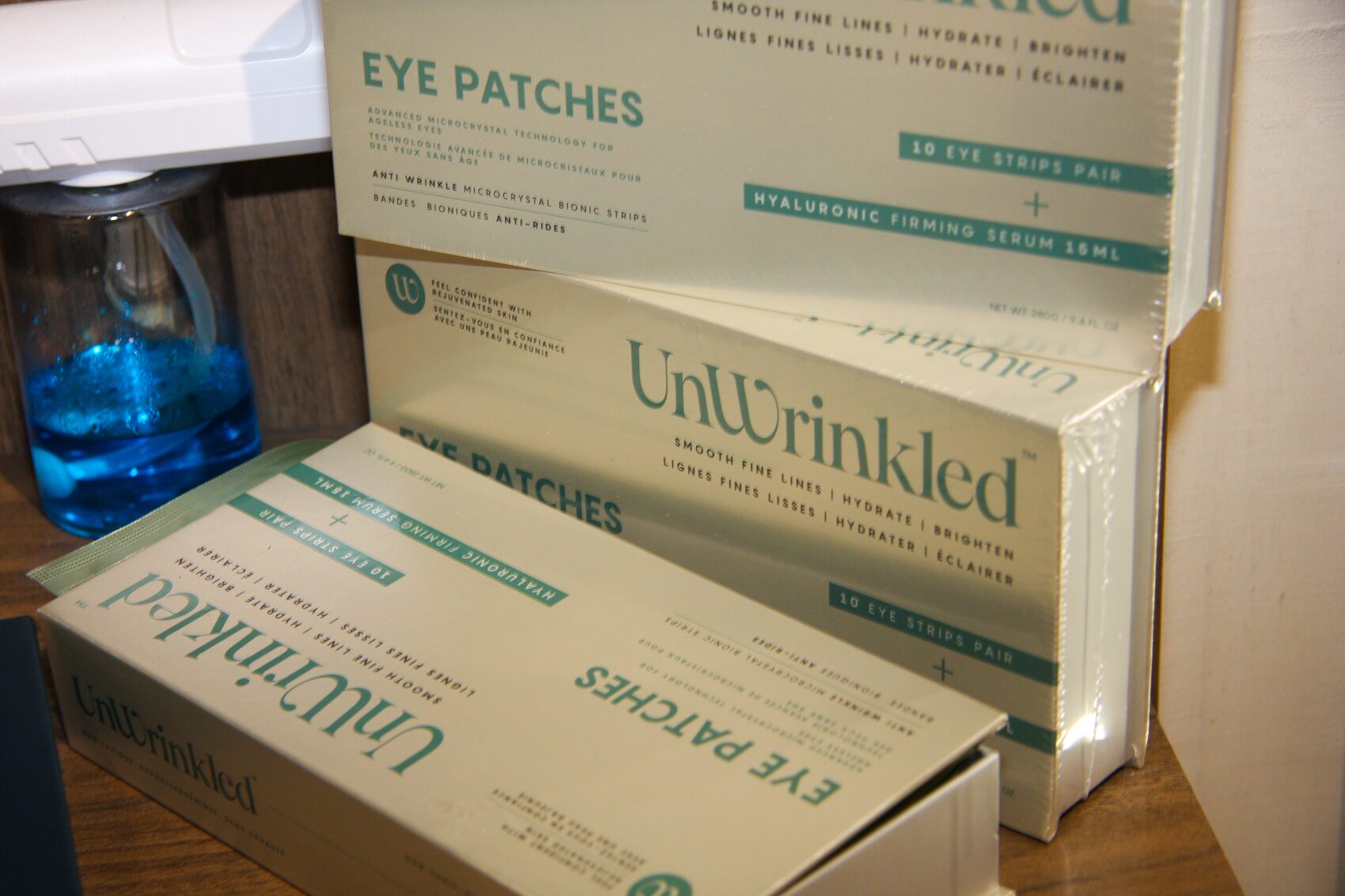 Three stacked UnWrinkled Eye Patches cartons on a dark wood surface, lit from the left. The packaging copy is legible: Advanced microcrystal technology for ageless eyes, anti wrinkle microcrystal bionic strips, smooth fine lines, hydrate, brighten, in English and French, with 10 eye strips pair plus hyaluronic firming serum 15ml.