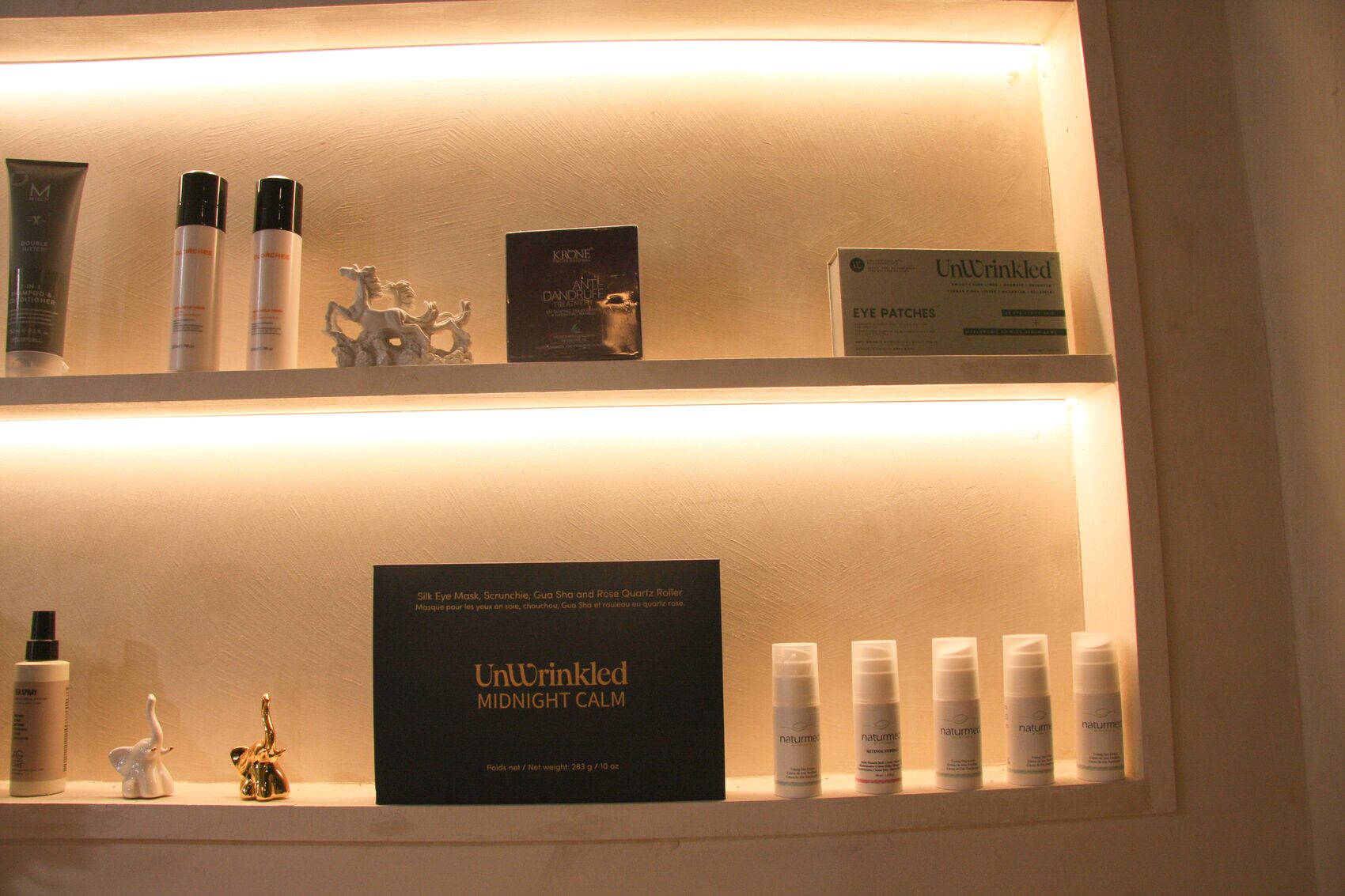 Two backlit shelves in a salon retail niche. On the lower shelf a navy UnWrinkled Midnight Calm box reading Silk Eye Mask, Scrunchie, Gua Sha and Rose Quartz Roller, with five white serum pumps ranged to its right and two small ceramic elephants to its left; on the upper shelf haircare tubes, a dandruff treatment carton and an UnWrinkled Eye Patches box.