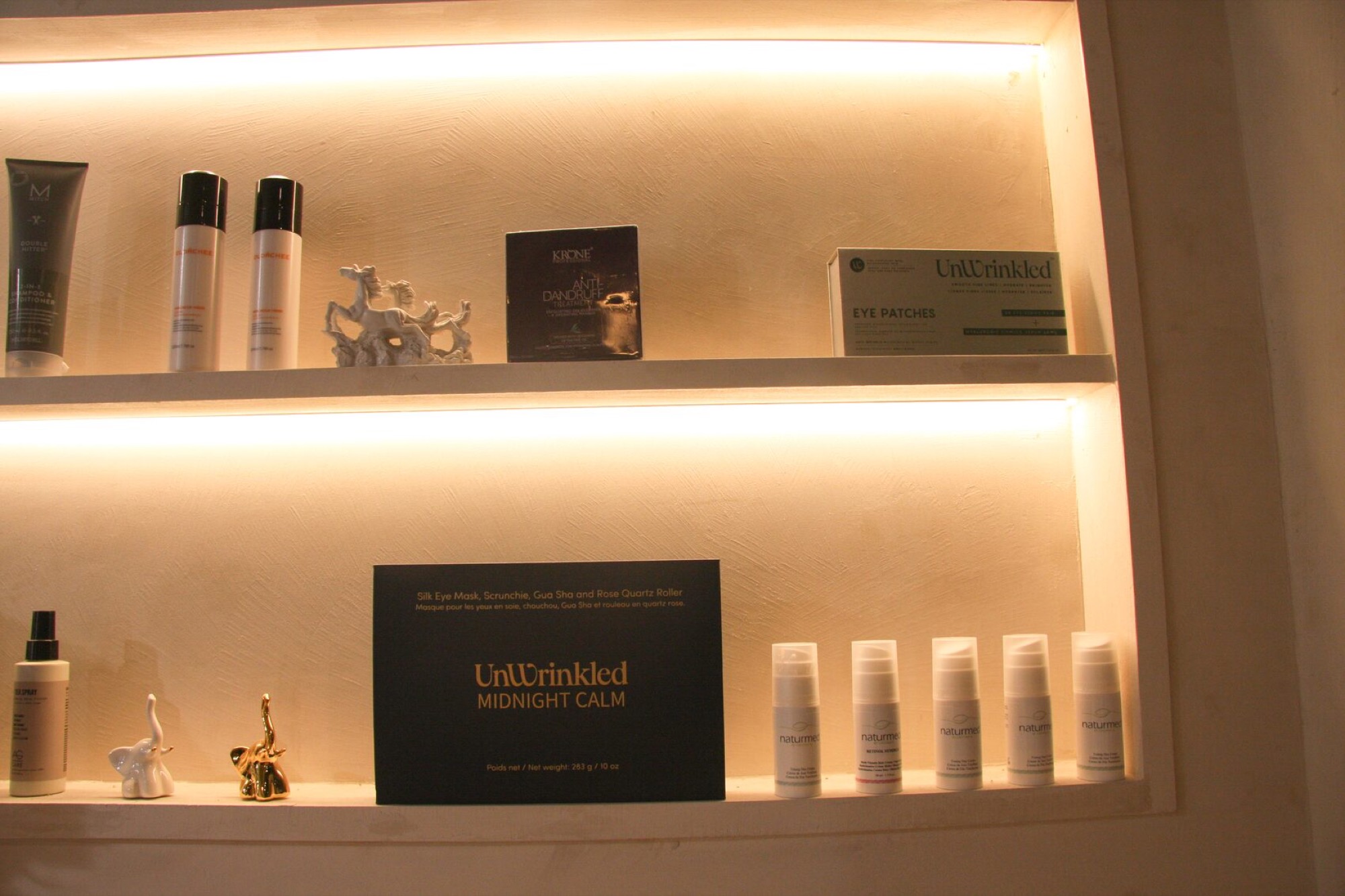 Two warm-lit retail niches: on the lower shelf a black UnWrinkled Midnight Calm box reading Silk Eye Mask, Scrunchie, Gua Sha and Rose Quartz Roller in English and French, net weight 283 g, flanked by a white and a gold elephant ornament and five naturmed pumps; an UnWrinkled Eye Patches carton sits on the shelf above.