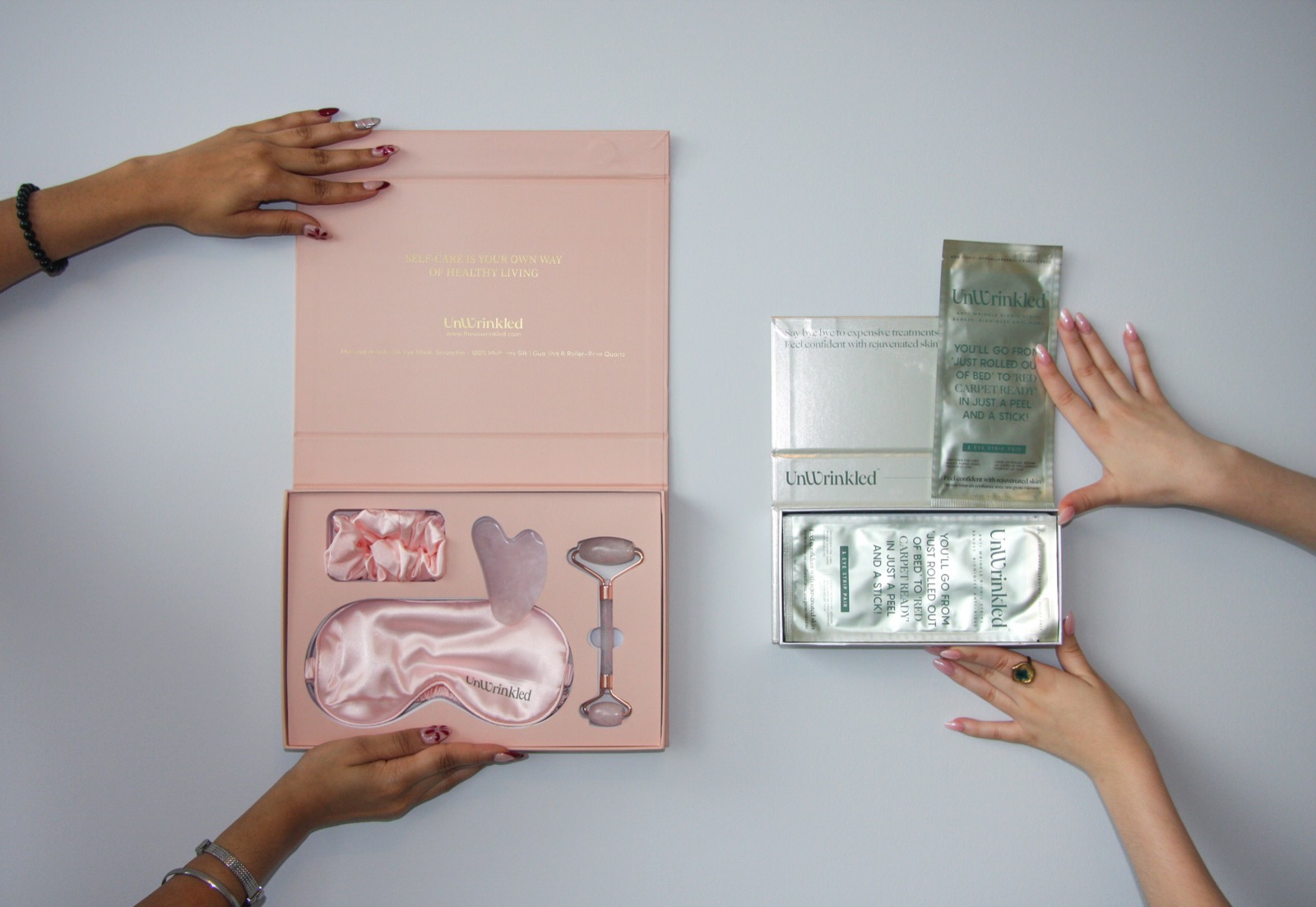 An overhead flat-lay on pale grey: an open pink Unwrinkled gift box holding a scrunchie, a gua sha stone, a rose-quartz roller and a silk sleep mask, beside a silver eye-strip carton and loose sachets, with four hands arranging them.