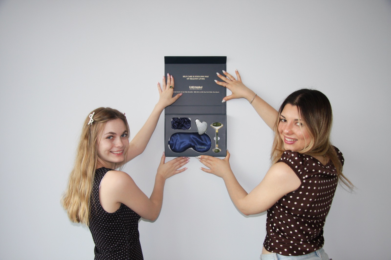 Two models against a lit studio wall holding an open navy UnWrinkled gift box towards camera.