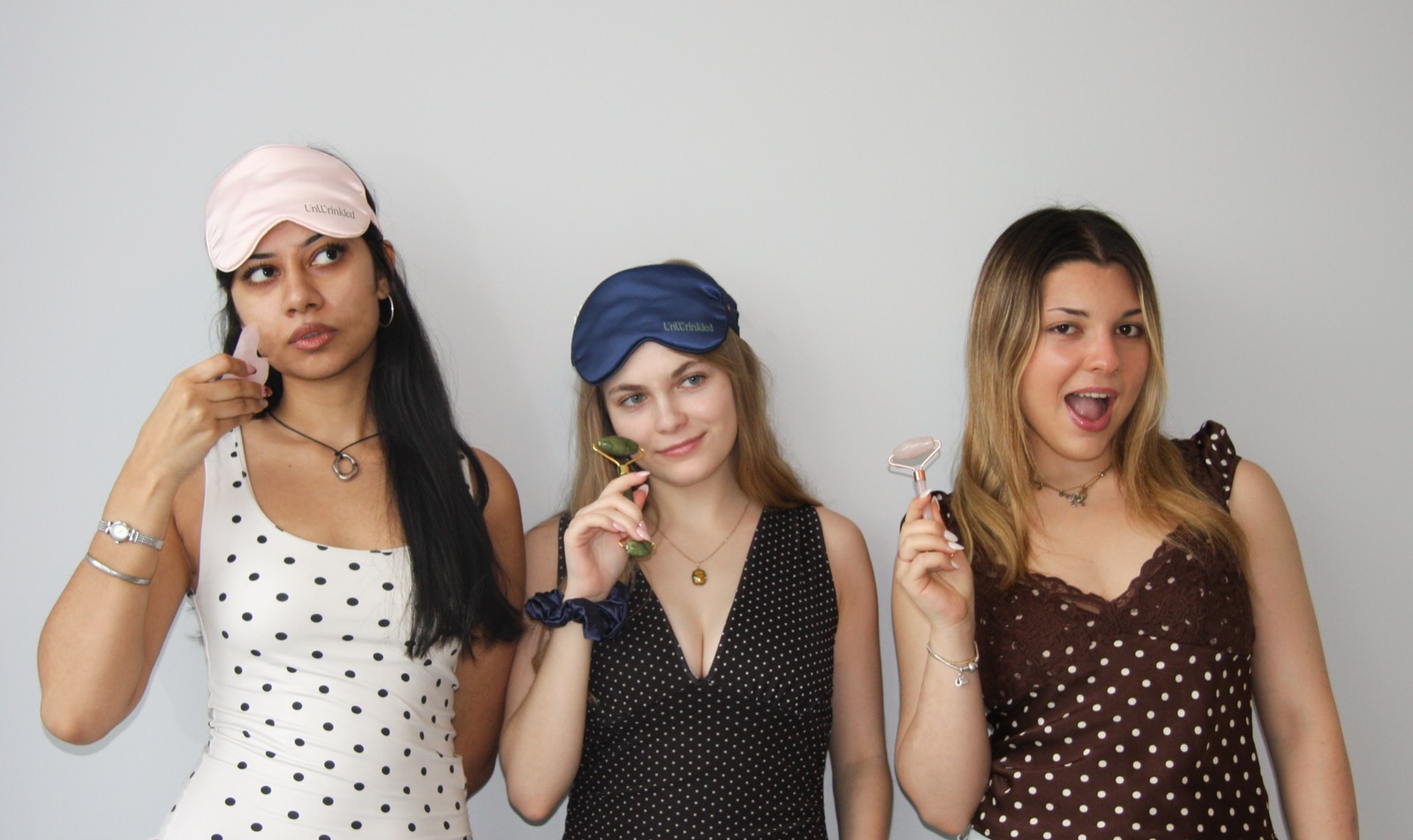 Three women side by side on a grey studio backdrop, each holding an Unwrinkled tool — a jade roller, a gua sha stone and a sheet mask.