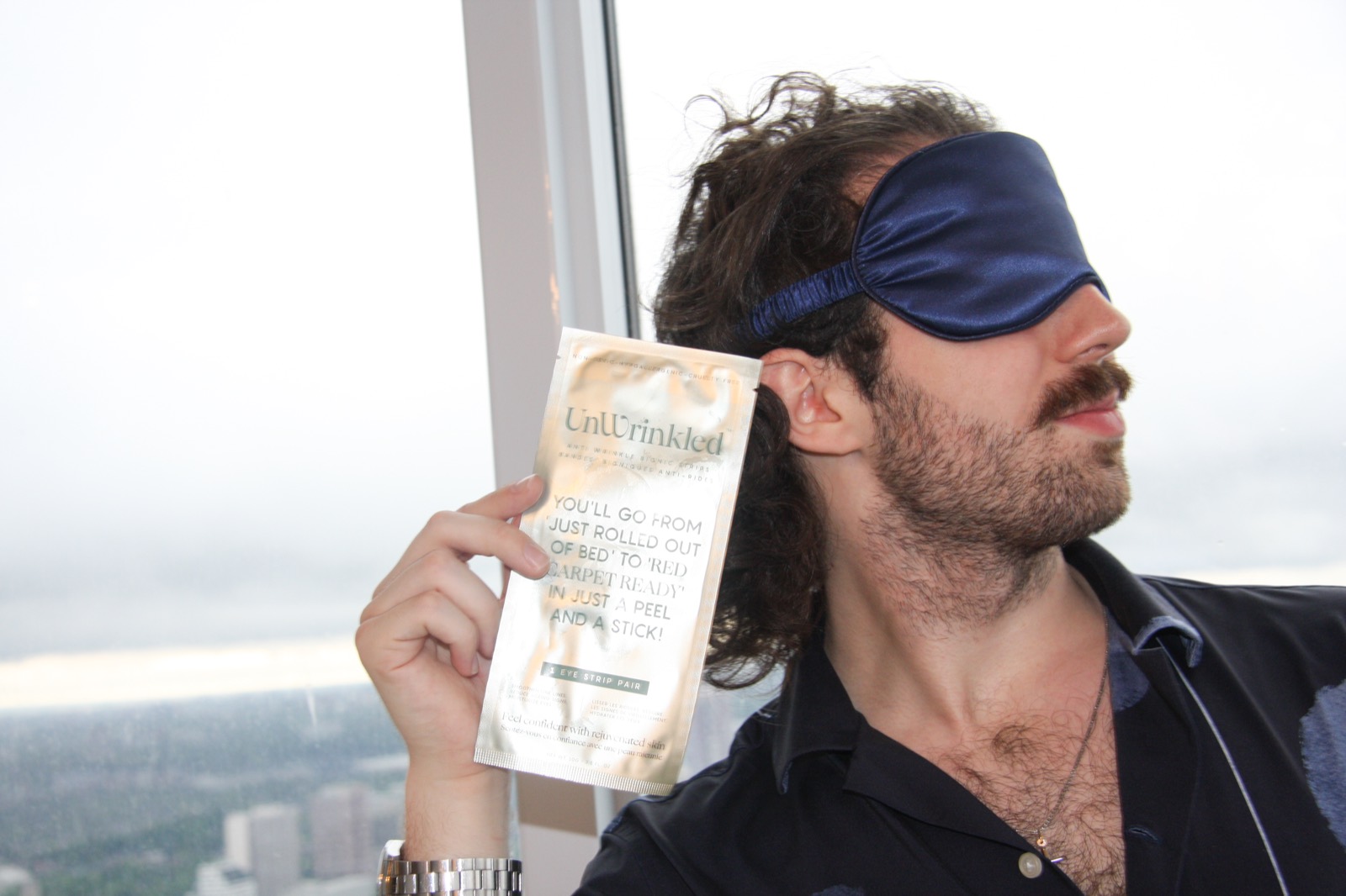 A model in a navy UnWrinkled silk eye mask holding an eye-strip sachet to camera, lit by a window with the Toronto skyline behind.
