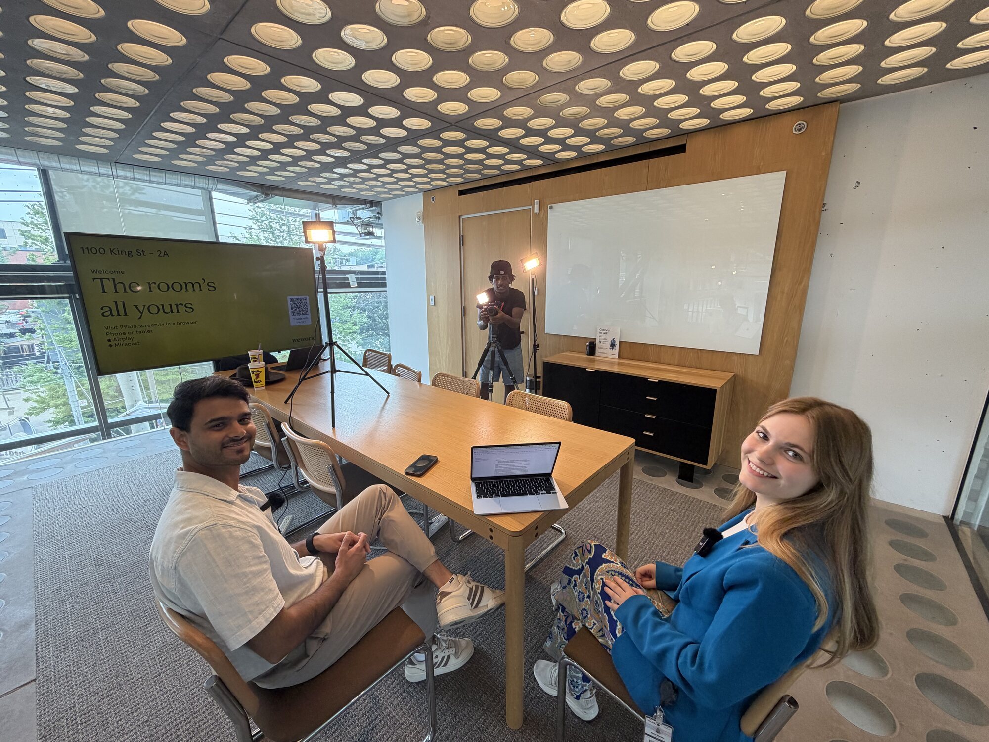 A TNAADO interview built in a client meeting room: two subjects seated at the table on lavalier microphones, an LED panel on a stand, and the operator shooting from a tripod at the far end.