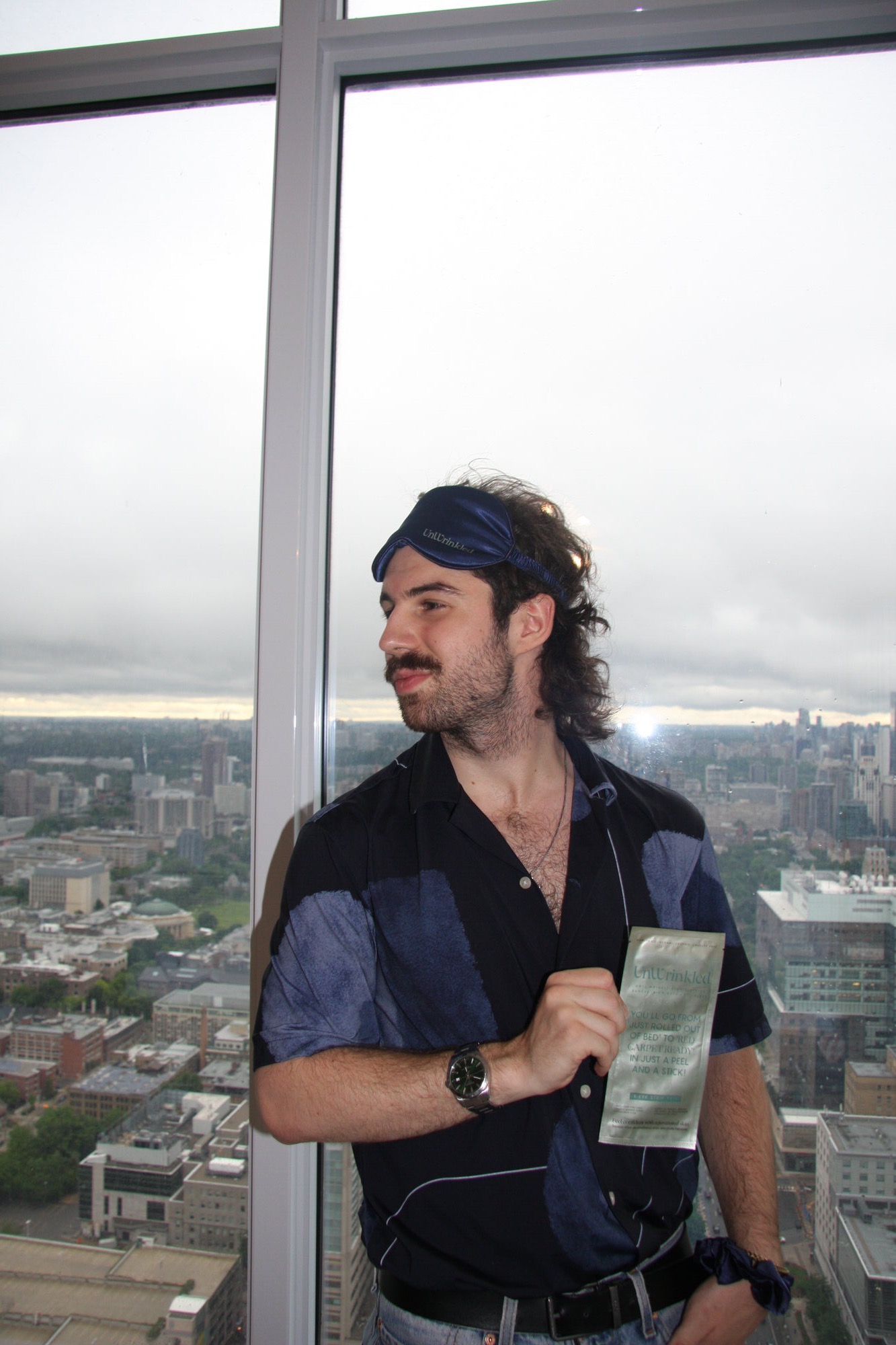 TNAADO's founder modelling for the UnWrinkled shoot, wearing the branded silk eye mask pushed back on his head and holding an eye-strip sachet to camera, with the Toronto skyline through the window behind him.