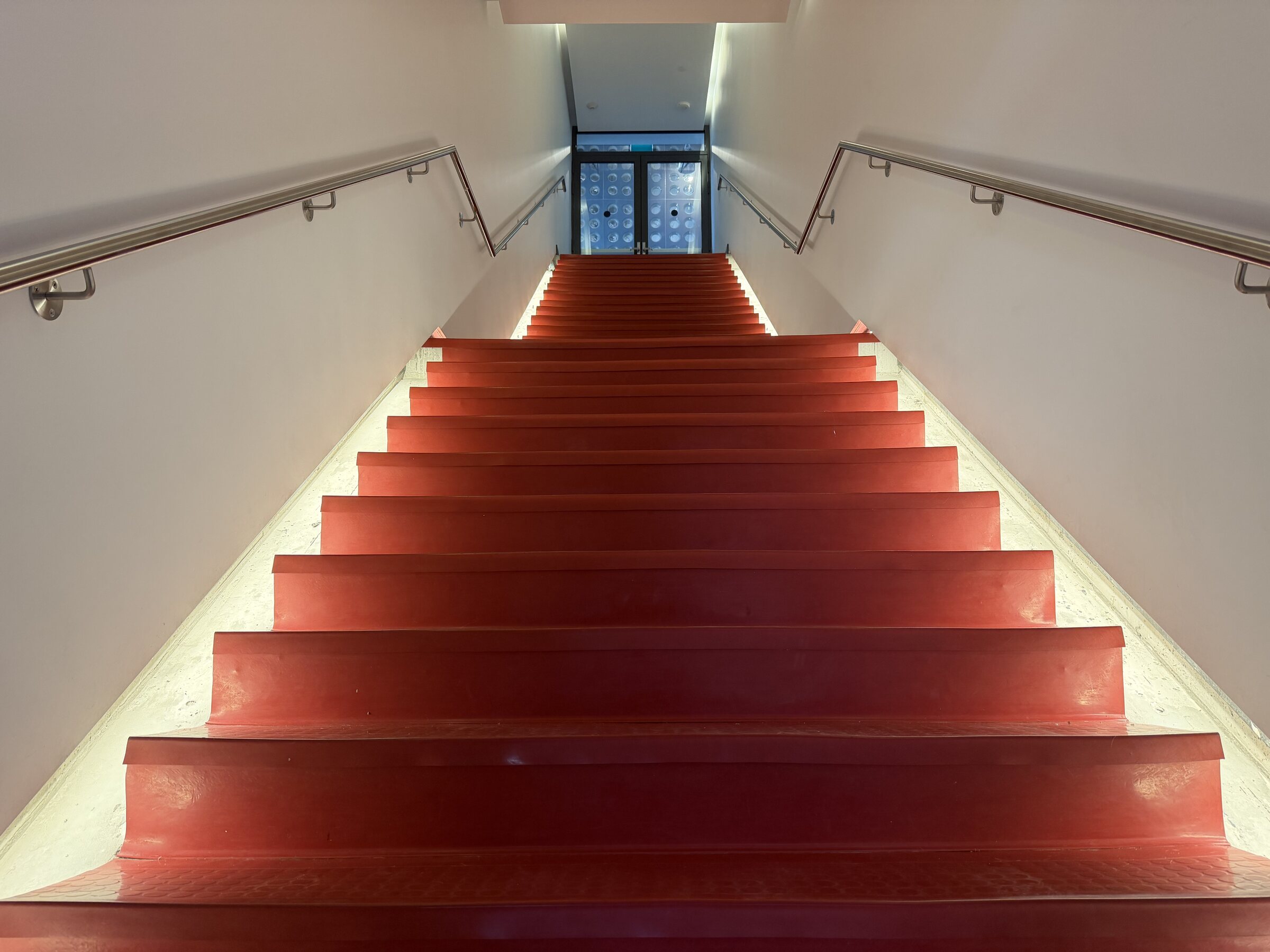 The staircase at TNAADO&rsquo;s Toronto office: deep red treads rising between white walls toward a lit glass doorway at the top.
