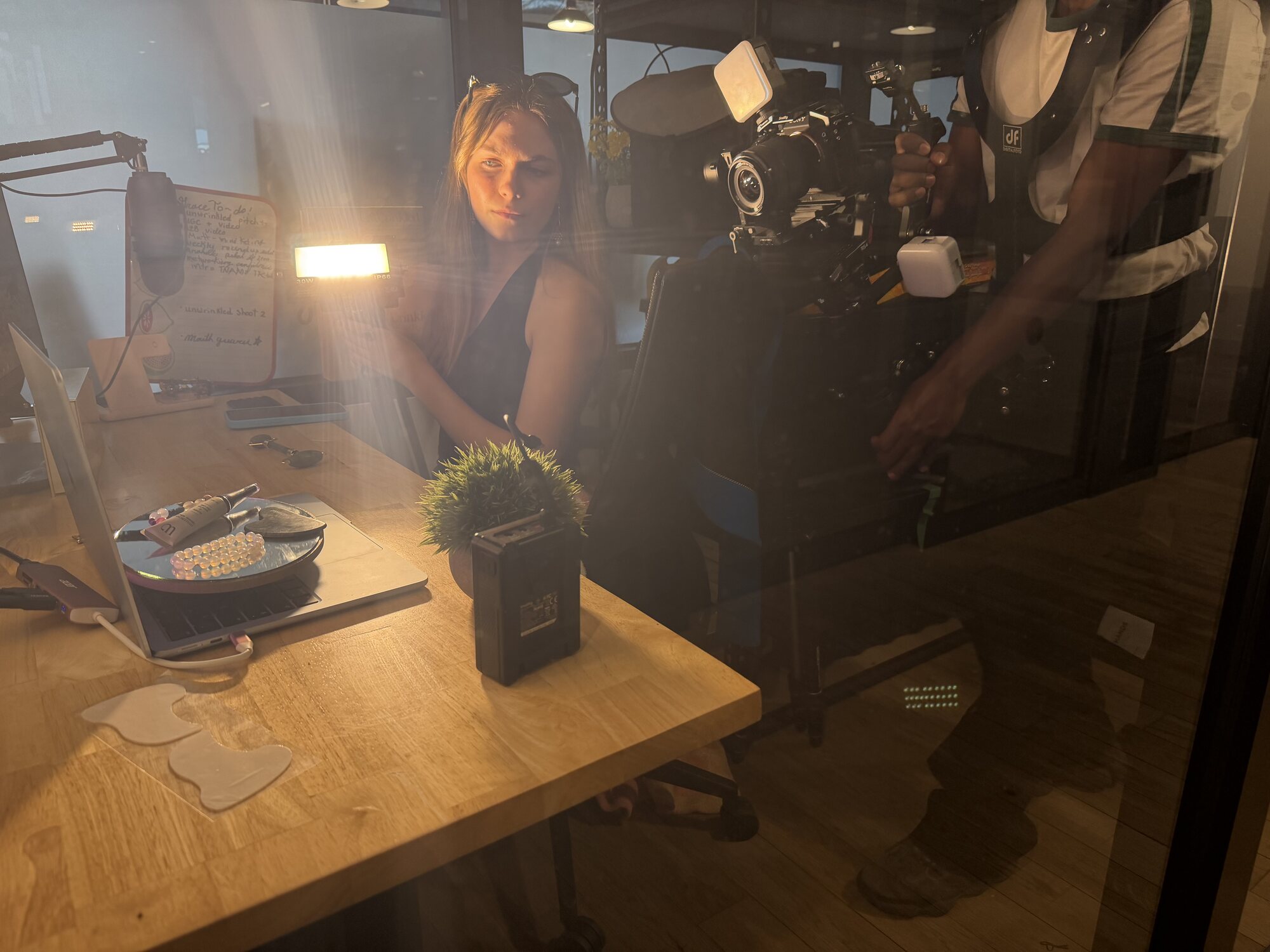 A TNAADO operator in a body-support vest running a Sony camera on a gimbal with an on-camera LED, shooting a product video across a lit desk where a producer is working.
