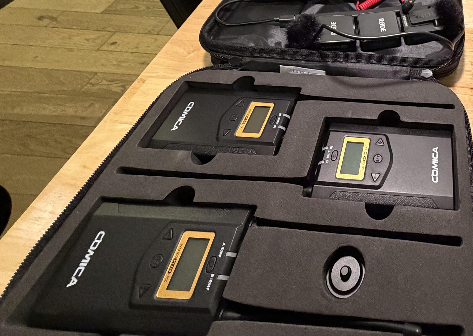 An open audio case on a desk: three lavalier transmitter packs seated in cut foam, with a wireless receiver and a fur windshield behind them.
