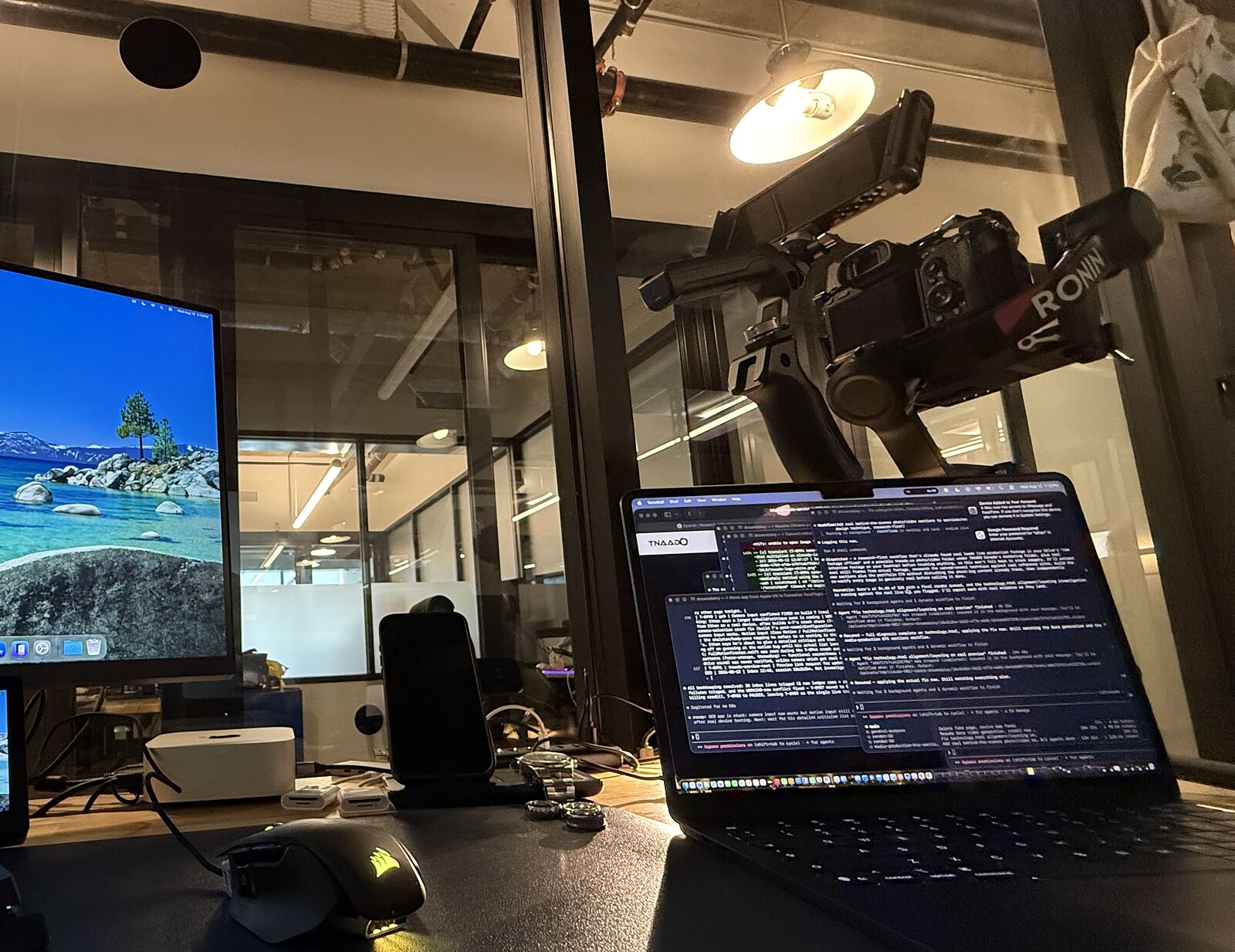 A TNAADO edit bay at night: a camera rigged above the desk on a gimbal arm, an external reference display, a Mac desktop and an open laptop mid-edit.